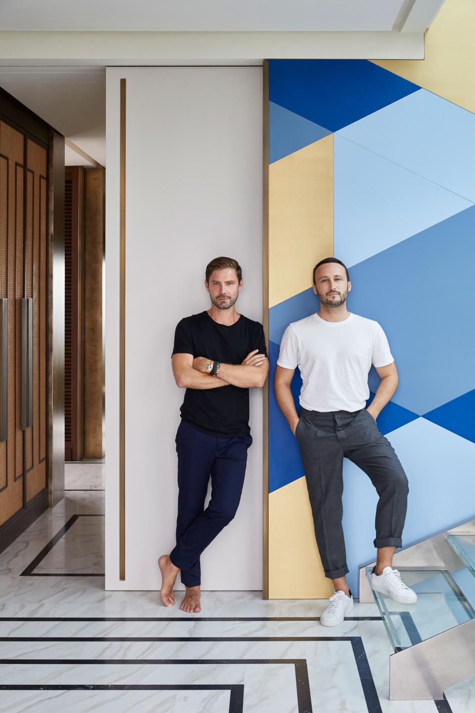 Emil Humbert (left) and Christophe Poyet standing in front of the wall composed of wood panels in brass and three shades of blue—Marine, Azur, and French. The floor is Calacatta Sponda and Nero Marquina; the cupboards in the background are caning, stained oak, and bronze handles.