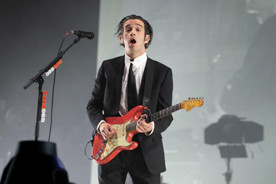 Matty Healy with a guitar wearing a suit and tie performing on stage