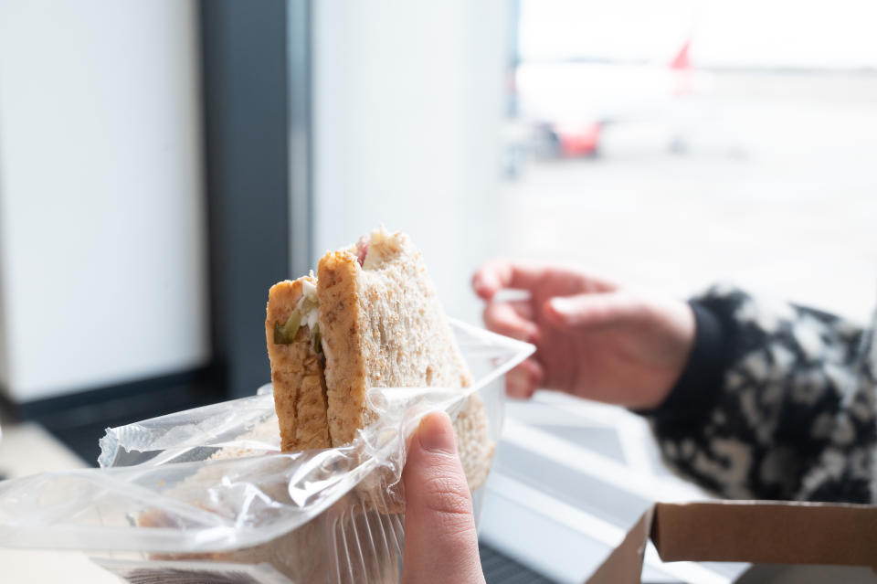 Person holding a packed sandwich near an airport window, with an airplane visible outside