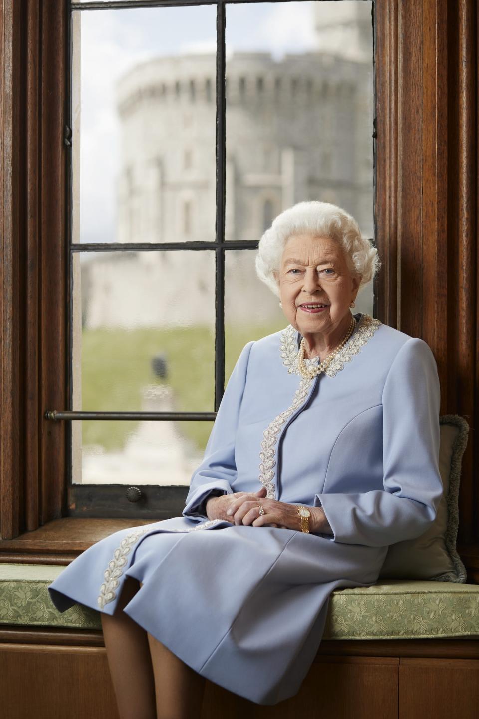 The Queen smiling and sitting on a small bench in front of a large window. She's wearing a pale blue coatdress with a white and silver petal design around her collar and down the center opening.