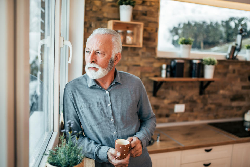 Portrait of a senior man standing in the kitchen and looking through window, holding a cup of coffee or tea on winter day.