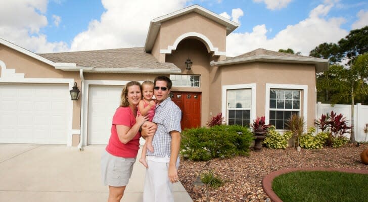 Image shows two adults with their baby in front of a large beige house with a landscaped front yard and spacious driveway. SmartAsset analyzed IRS data to conduct its study on where upper-middle-class people are moving.