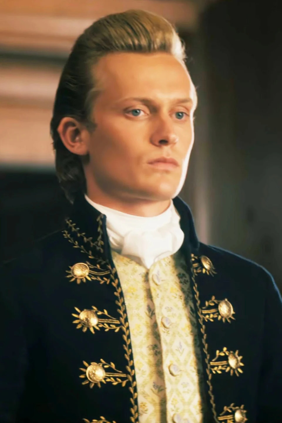 Freddie Dennis as Reynolds, wears a period costume with intricate embroidery and a high collar, standing in an elegant setting