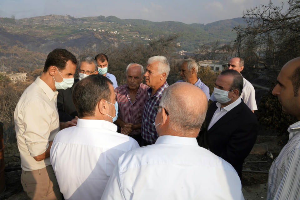 In this photo released Tuesday, Oct. 13, 2020 on the official Facebook page of the Syrian Presidency, Syrian President Bashar Assad, left, wearing a mask to help prevent the spread of the coronavirus, speaks with people during his visit to the coastal province of Latakia, Syria. Assad made a rare public visit to Latakia where he toured areas that suffered heavy damage in last week’s deadly wildfires. (Syrian Presidency via Facebook via AP)