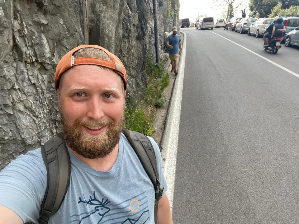 Timothy, wearing a backwards orange hat, blue shirt, and backpack, walks along a busy narrow road with his husband following behind.