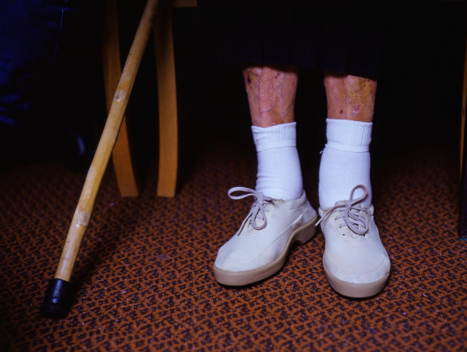 Person standing with white shoes and socks, cane to the left, on patterned carpet