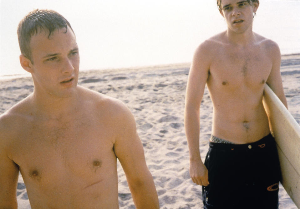 Still from the movie "Bully" showing two men (actors Brad Renfro and Nick Stahl) walking topless on a beach, one of them carrying a surfboard