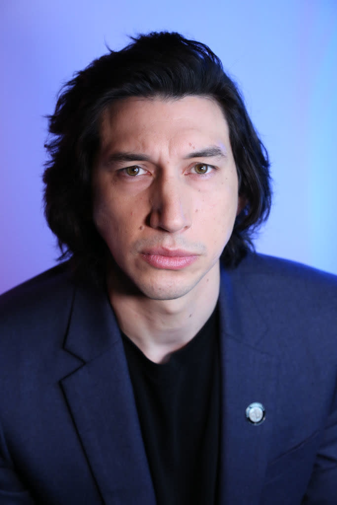 Adam Driver is wearing a blazer and a black shirt while looking directly at the camera. The background has a soft gradient