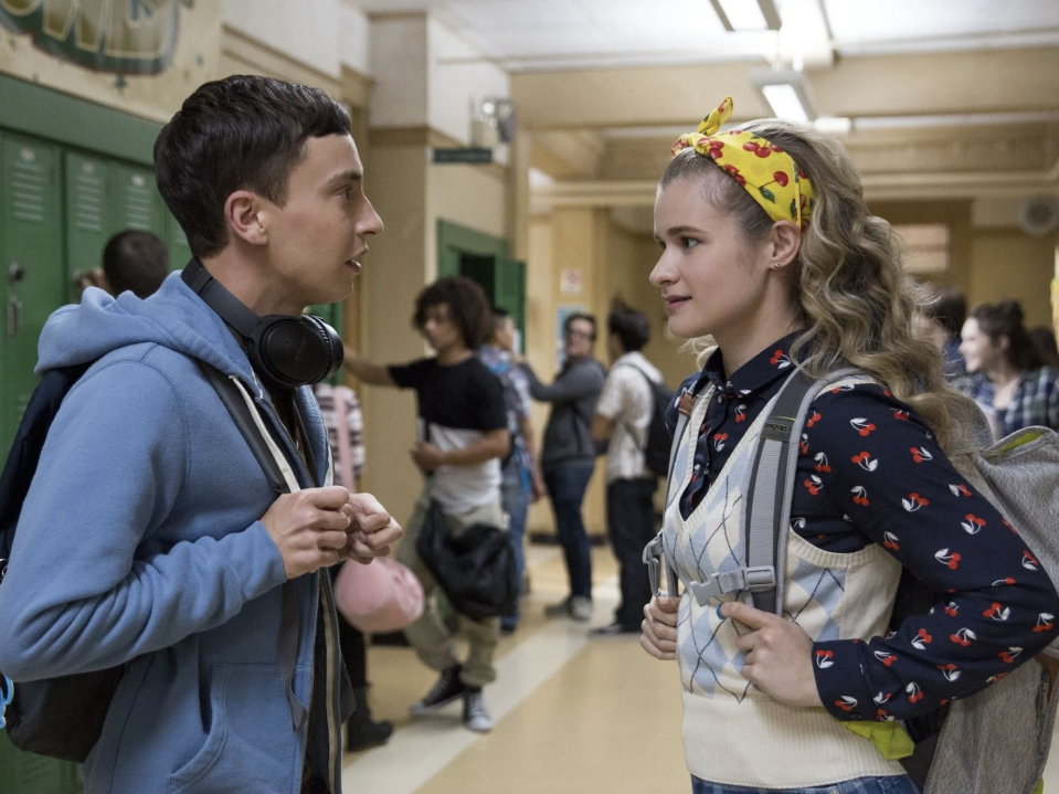 A young man and woman are talking in a busy school hallway. The man wears a hoodie and has headphones around his neck. The woman wears a headband and a sweater