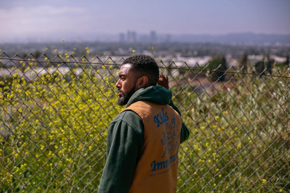 A man in a hoodie holds onto a chain link fence in front of a field of flowers.