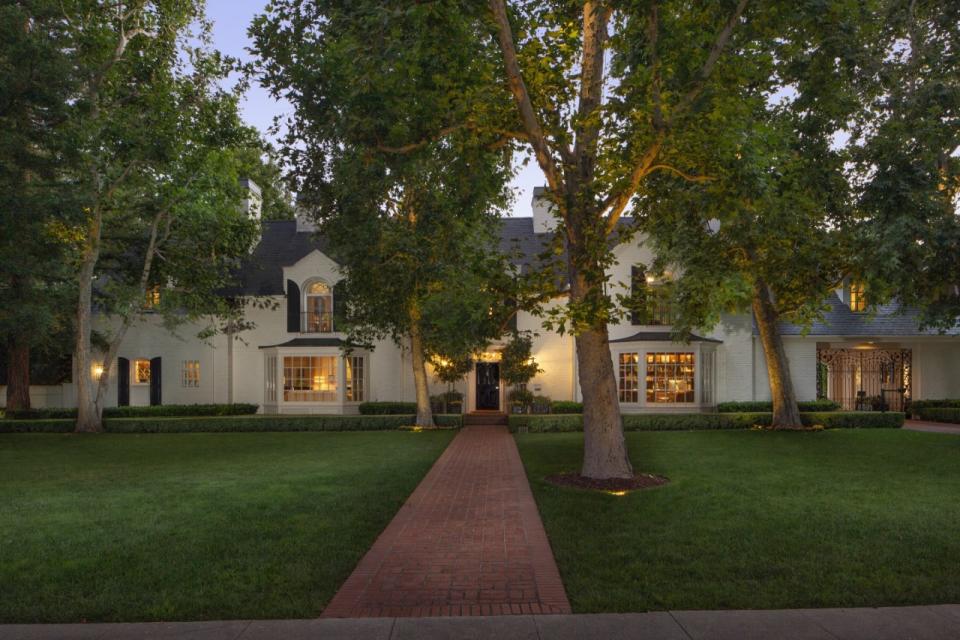 A brick walkway leads past mature trees to the two-story home's front door, bordered by bay windows.