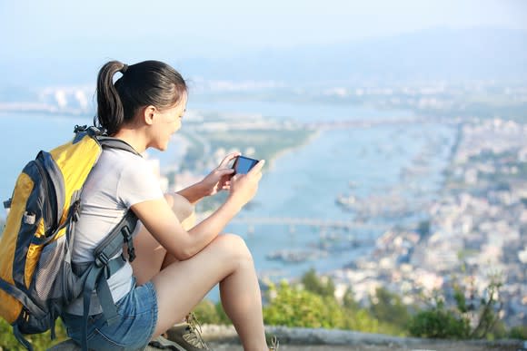 Asian woman uses a smartphone while sitting on a hill overlooking a Chinese fishing village.