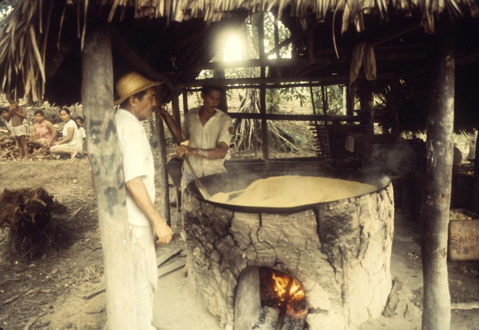 man stands next to a large barrel with fire underneath, under a thatched roof