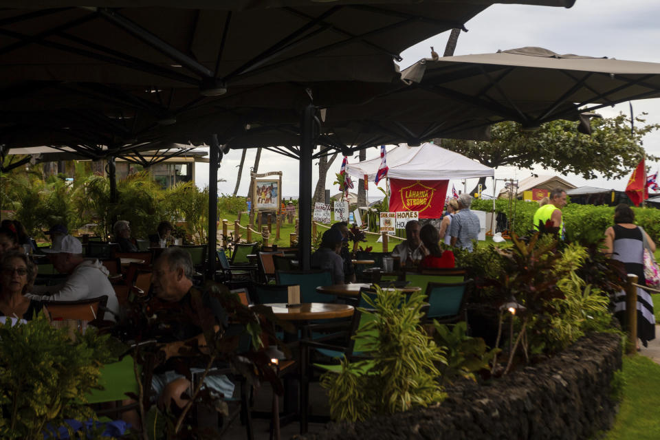 Tourists visit at a outdoor restaurant next to the Lahaina Strong "Fish-in" on Friday, Dec. 1, 2023, in Lahaina, Hawaii. Lahaina Strong has set up a "Fish-in" to protest living accommodations for those displaced by the Aug. 8, 2023 wildfire, the deadliest U.S. wildfire in more than a century. More than four months after the fire, tensions are growing between those who want to welcome tourists back to provide jobs and those who feel the town isn't ready for a return to tourism. (AP Photo/Ty O'Neil)