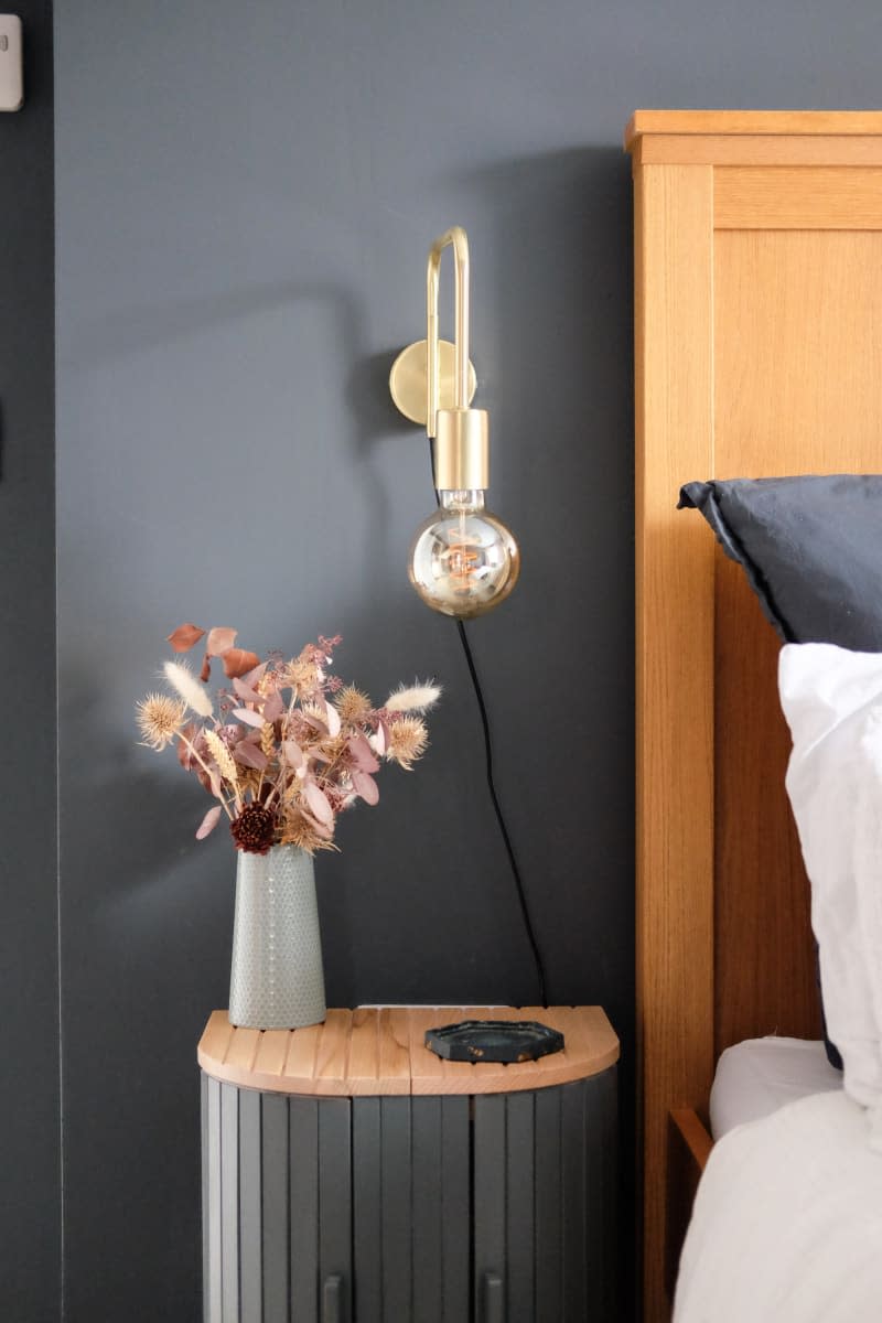 slate gray walls with light wood headboard and gray and wood slated end table, gold sconce fixture and white linens