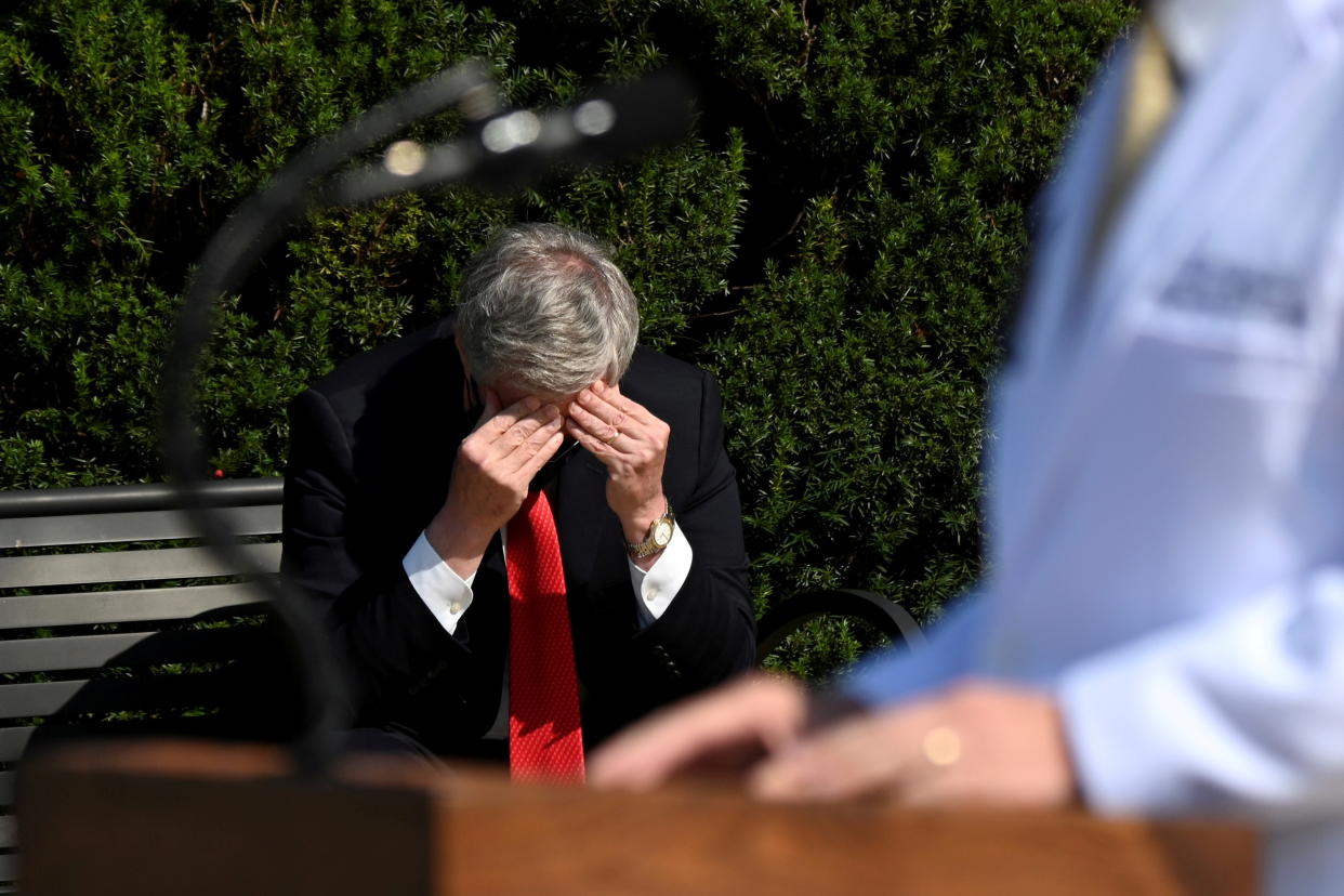 Mark Meadows, sitting on a bench, looks down and rubs his head as Dr. Sean Conley speaks at a podium about President Trump’s condition outside Walter Reed hospital on October 4, 2020.