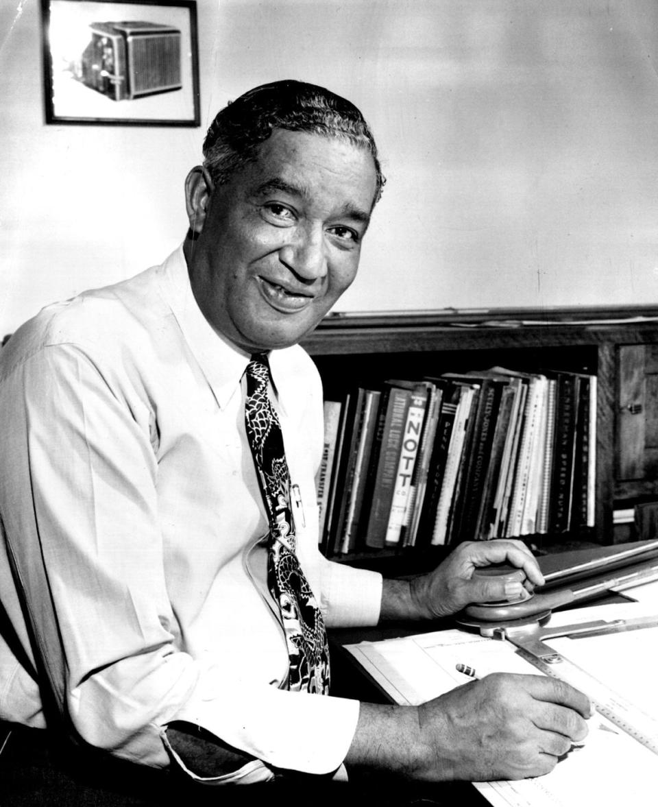 frederick mckinley jones smiles at the camera while sitting at a table with a pencil in hand, paper and equipment rests on the table in front of him, behind is a bookshelf, he wears a collared shirt and patterned tie