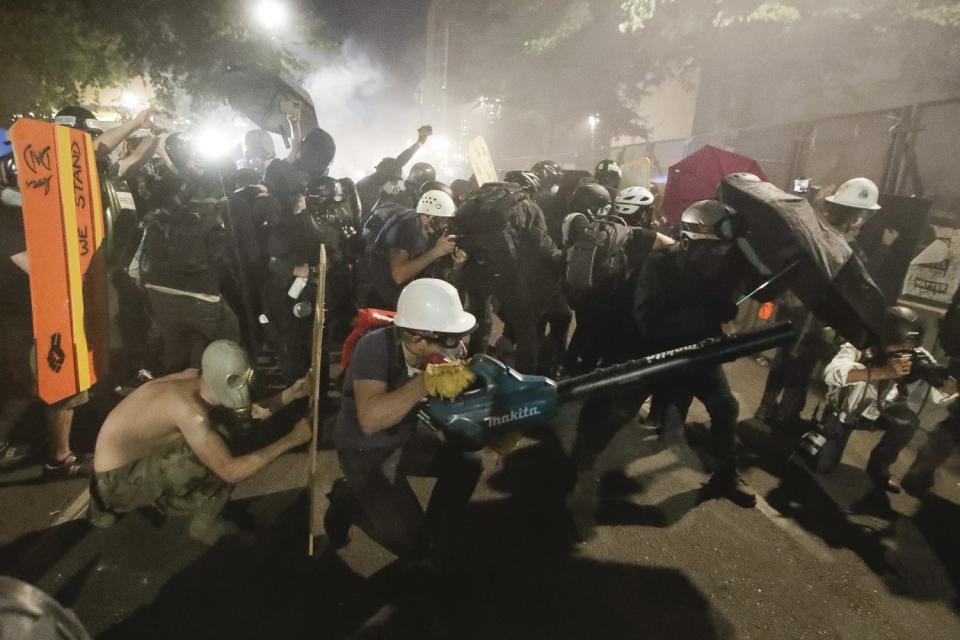 Demonstrators huddle behind shields and umbrellas as one uses a leaf blower to blow back tear gas fired by federal agents