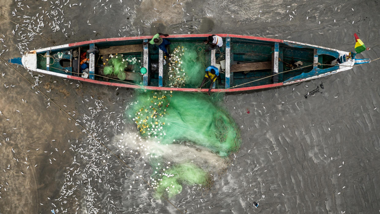 An aerial shot of a fishing boat with three people aboard and many visible fish in the clear water.