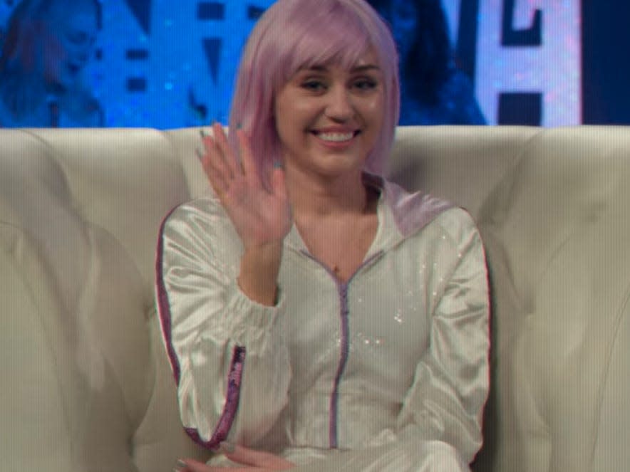 miley cyrus as ashley o on black mirror, wearing a shiny white tracksuit and pink wig, and waving, sitting in a white chair