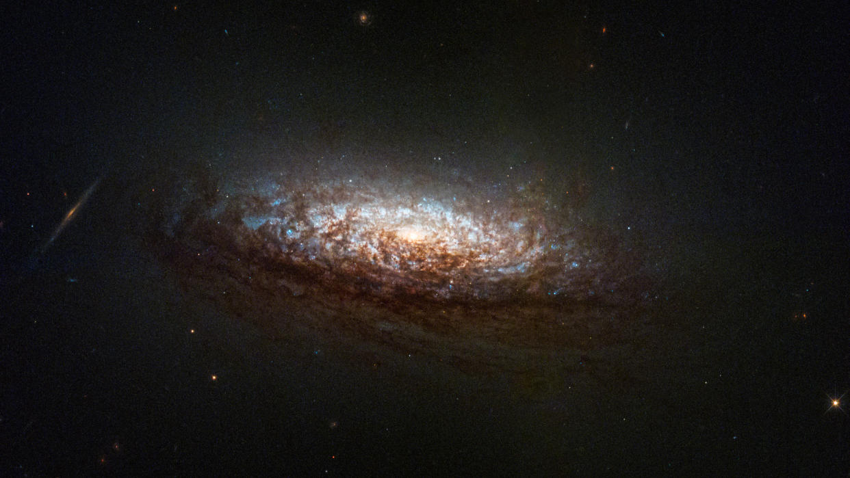  A stunning spiral galaxy hangs dimly in the dark of space; a bright light at its center illuminates nearby gasses, swirling around in shades of pale blues, whites and crisp browns. 