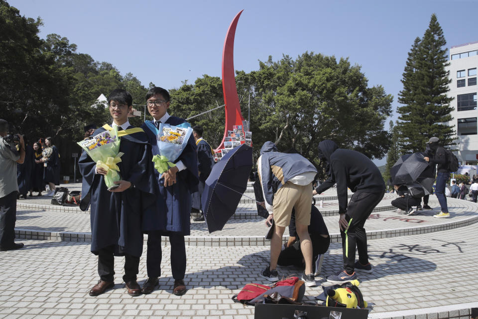 Graduates stand near graffiti against police brutality at a graduation ceremony that was disrupted by protesters at a the University of Science and Technology in Hong Kong on Friday, Nov. 8, 2019. The ceremony was cut short, and black-clad masked students turned the stage into a memorial for Chow Tsz-Lok who fell off a parking garage after police fired tear gas during clashes with anti-government protesters and died Friday. (AP Photo/Kin Cheung)