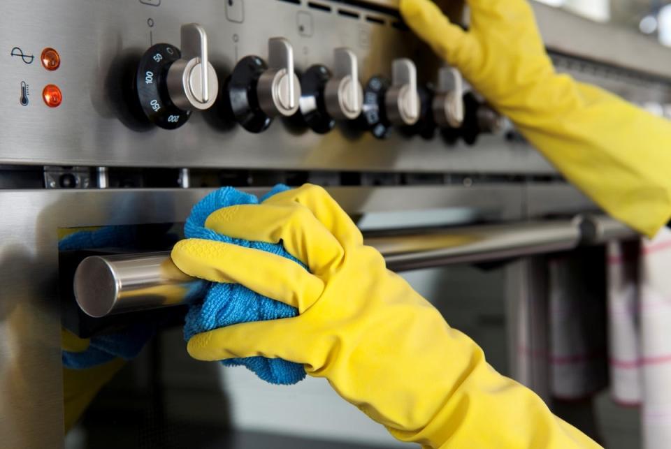 Yellow gloved hands cleanings a stainless steel stove with a blue microfiber cloth.