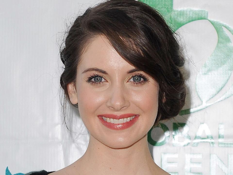 A headshot of Alison Brie smiling in front of a white and green background.