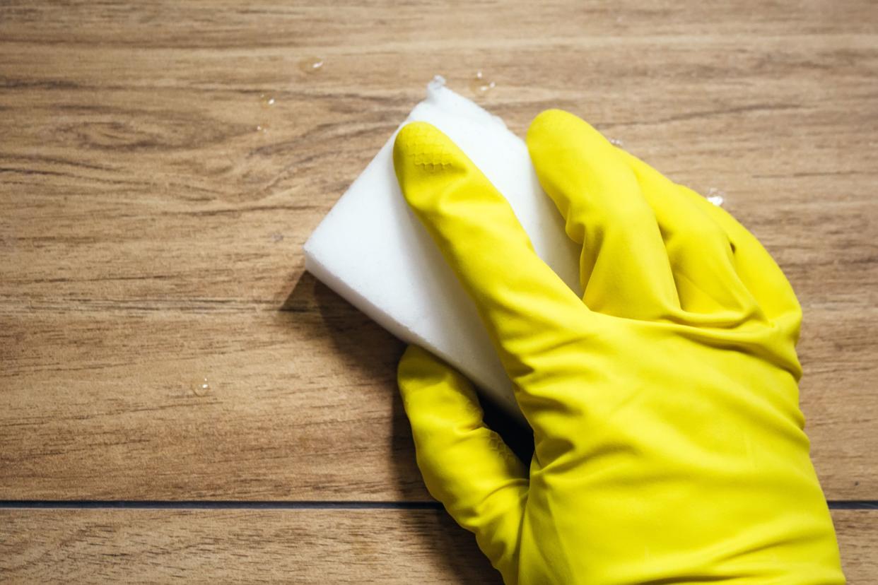 A hand in a yellow rubber glove wipes wet tile imitating a wooden surface with a white melamine sponge. Cleaning in a modern bathroom. Selective focus