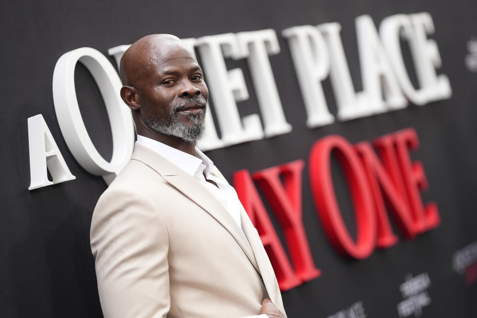 Djimon Hounsou on a red carpet for "A Quiet Place: Day One," wearing a stylish light-colored suit and white shirt, posing with a confident expression