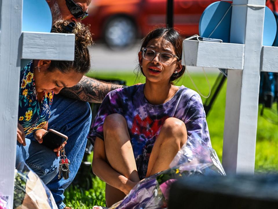 A girl sits in front of a cross holding her knees and crying.