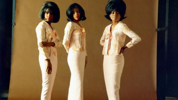 the supremes post for a photo on a honey colored background, they wear matching yellow blouses, white pants, and white high heels