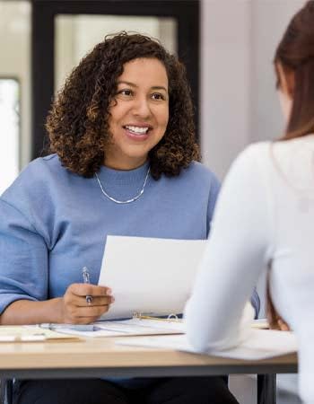 A woman smiles at a person sitting across from her while holding a pen a d document.