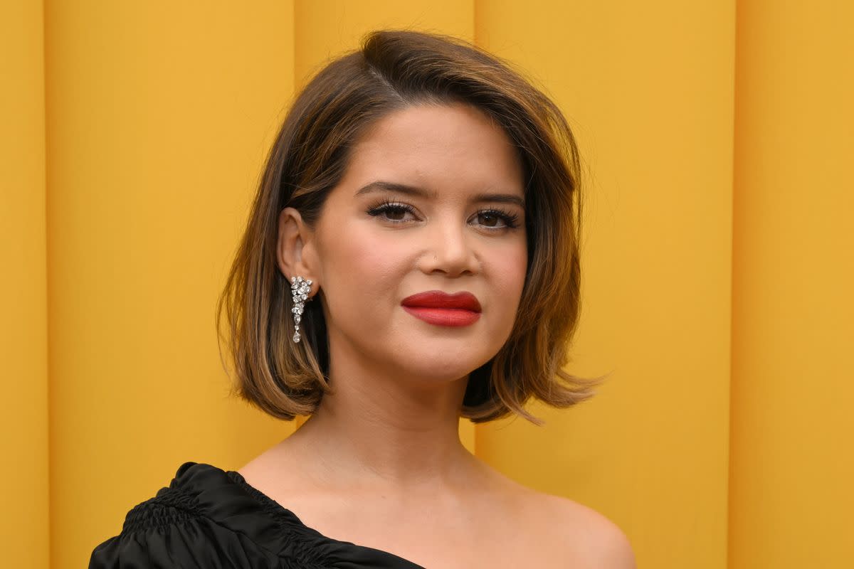 A white woman who has a short brunette bob and is wearing red lipstick stands in front of a yellow curtain. 