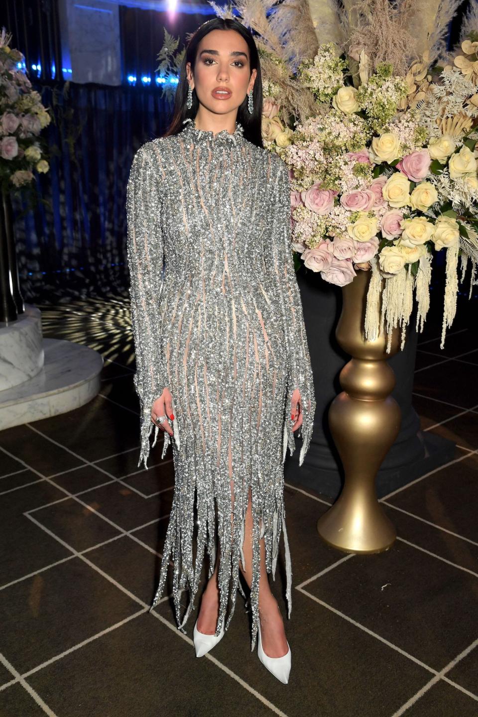 Dua Lipa stands in a silver, sparkly, see-through dress in front of flowers.