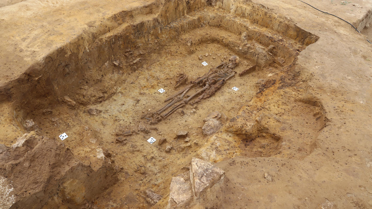  We see a large burial that's been excavated in a dirt area. There is a skeleton at the bottom. 