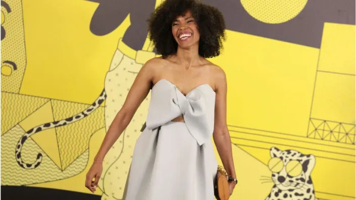 Lula Mebrahtu smiling in a beige dress with a yellow background behind her.