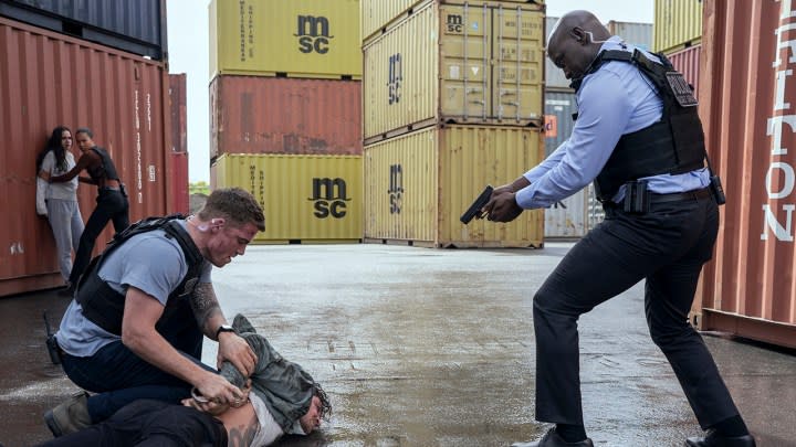 Peter holding down Colin, Agent Monks holding a gun to his, Maddie and Chelsea in the background of a shipping yard in a scene from The Night Agent.