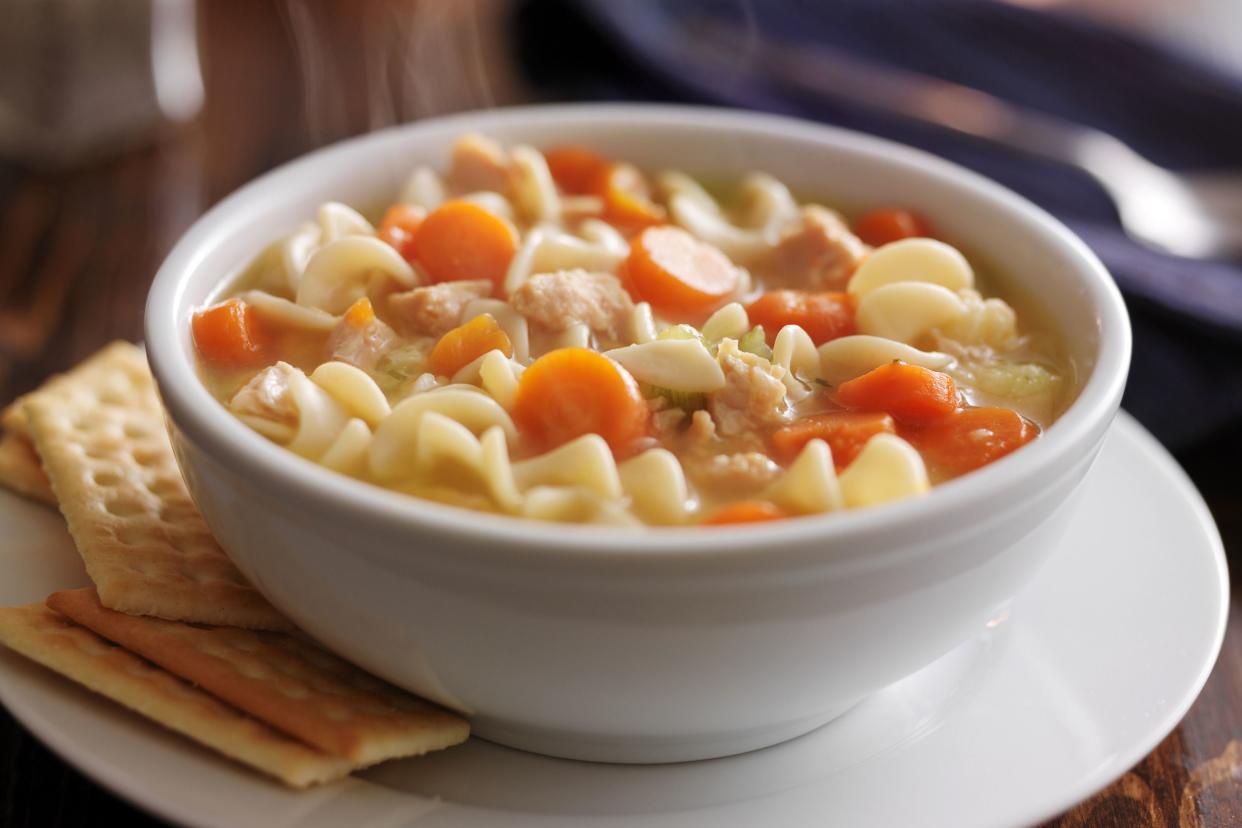 Closeup of slow cooker chicken noodle soup in a white porcelain bowl on a matching white plate, with a side of soda crackers on a wooden table with a blurred background