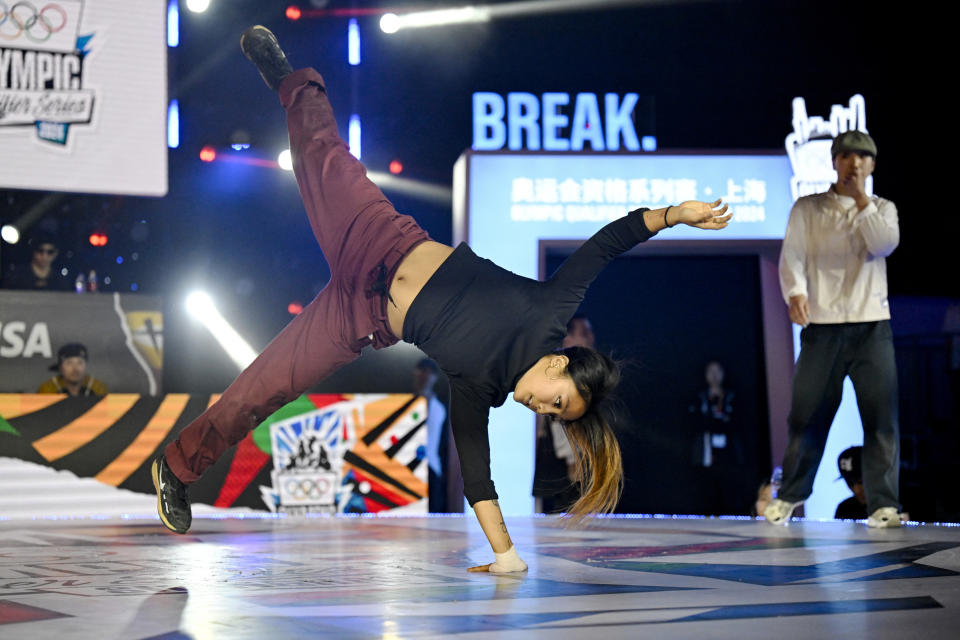 Breakdancer performing on one hand at an Olympic event, with another person watching in the background. The word "BREAK" is displayed behind them