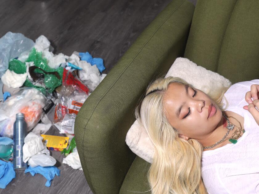 A person with long blonde hair lying on the edge of a couch with a pule of trash on the floor above them.