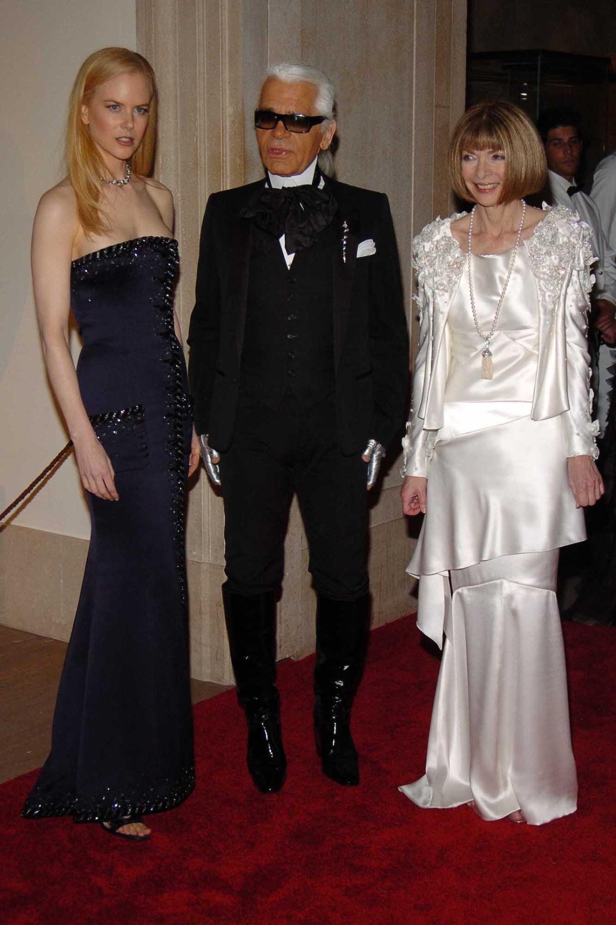 Nicole Kidman, Karl Lagerfeld, and Anna Wintour stand together at the 2005 Met Gala. Kidman is wearing a navy strapless dress, Lagerfeld a black suit with shiny knee-high boots, and Wintour a white silk dress and jacket.