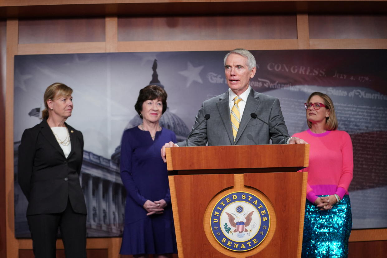 Senator Rob Portman stands at a podium. Behind him are Senators Tammy Baldwin, Susan Collins and Kyrsten Sinema.