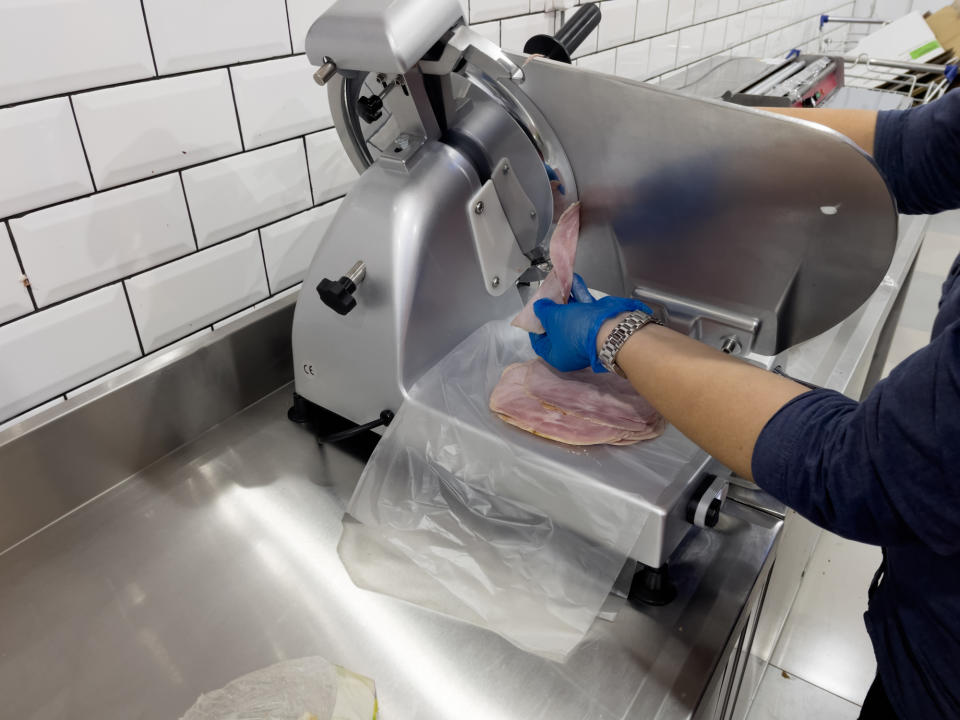 Person in a long-sleeve shirt and gloves slicing deli meat on a meat slicer in a commercial kitchen