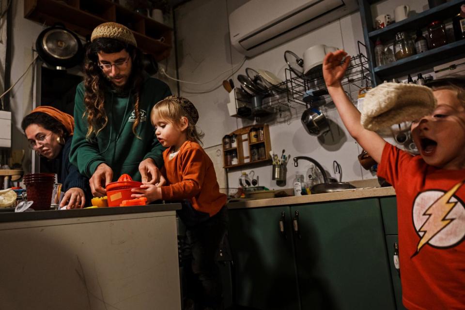 A man and woman preparing food in a kitchen as one small child helps and another plays nearby