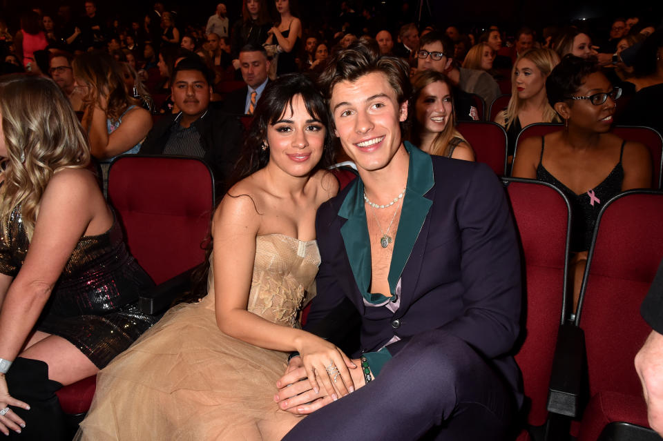 Camila Cabello in a strapless gown and Shawn Mendes in a dark suit with an open collar sitting close together at an event, smiling at the camera