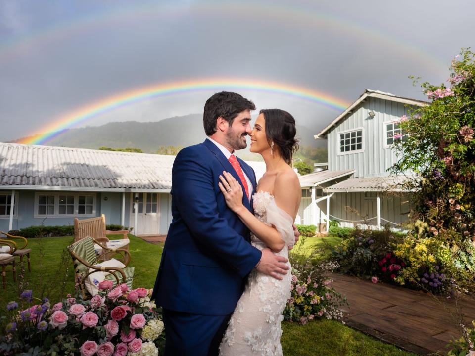 A bride and groom embrace with a rainbow behind them.