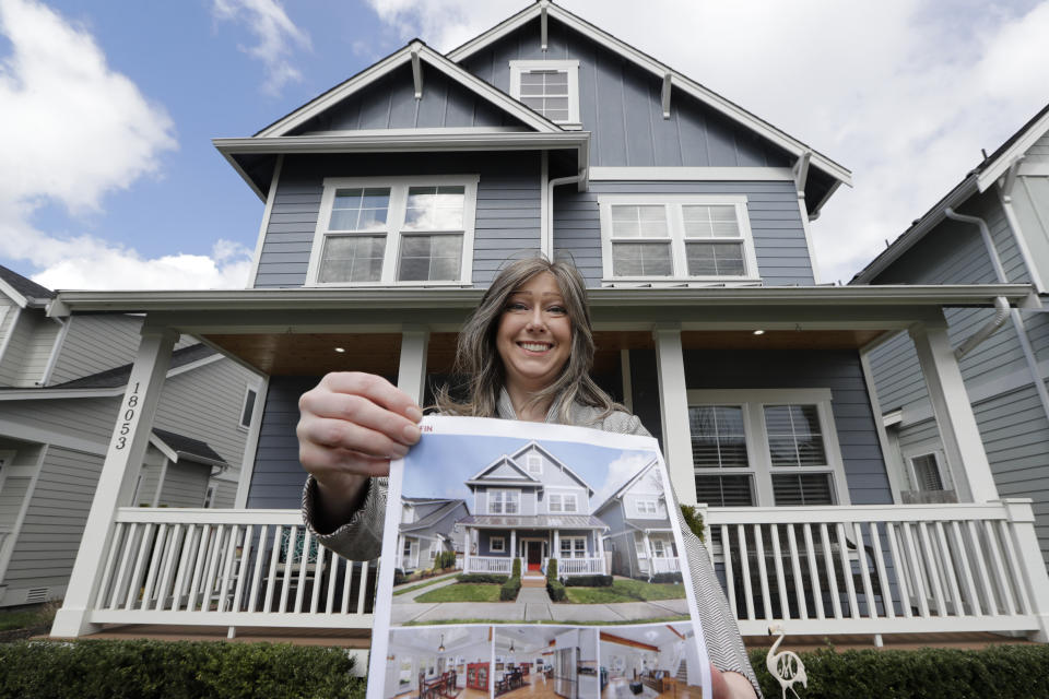 In this Wednesday, April 1, 2020 photo, Rebeka McBride holds a picture of her home as she poses in front of it in Monroe, Wash., outside of Seattle. She put her house on the market in early March and is in the process of closing on the sale. (AP Photo/Elaine Thompson)