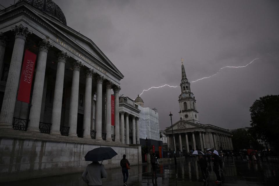 A lightning strike is pictured behind the church tower of St Martin-in-the-Fields as people walk past the National Portrait gallery under their umbrellas during a storm, in Trafalgar Square, in central London, on September 21, 2023. (Photo by JUSTIN TALLIS / AFP) (Photo by JUSTIN TALLIS/AFP via Getty Images)