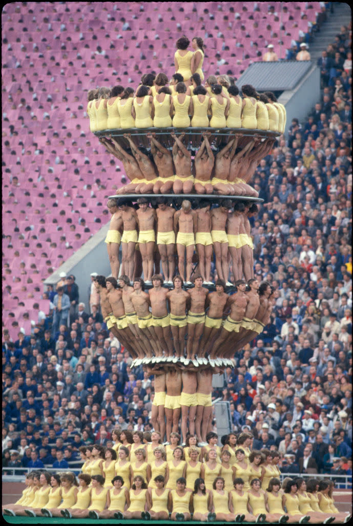 A large human torch of many levels, each one made up of male and female athletes; the base has three levels of people standing and sitting